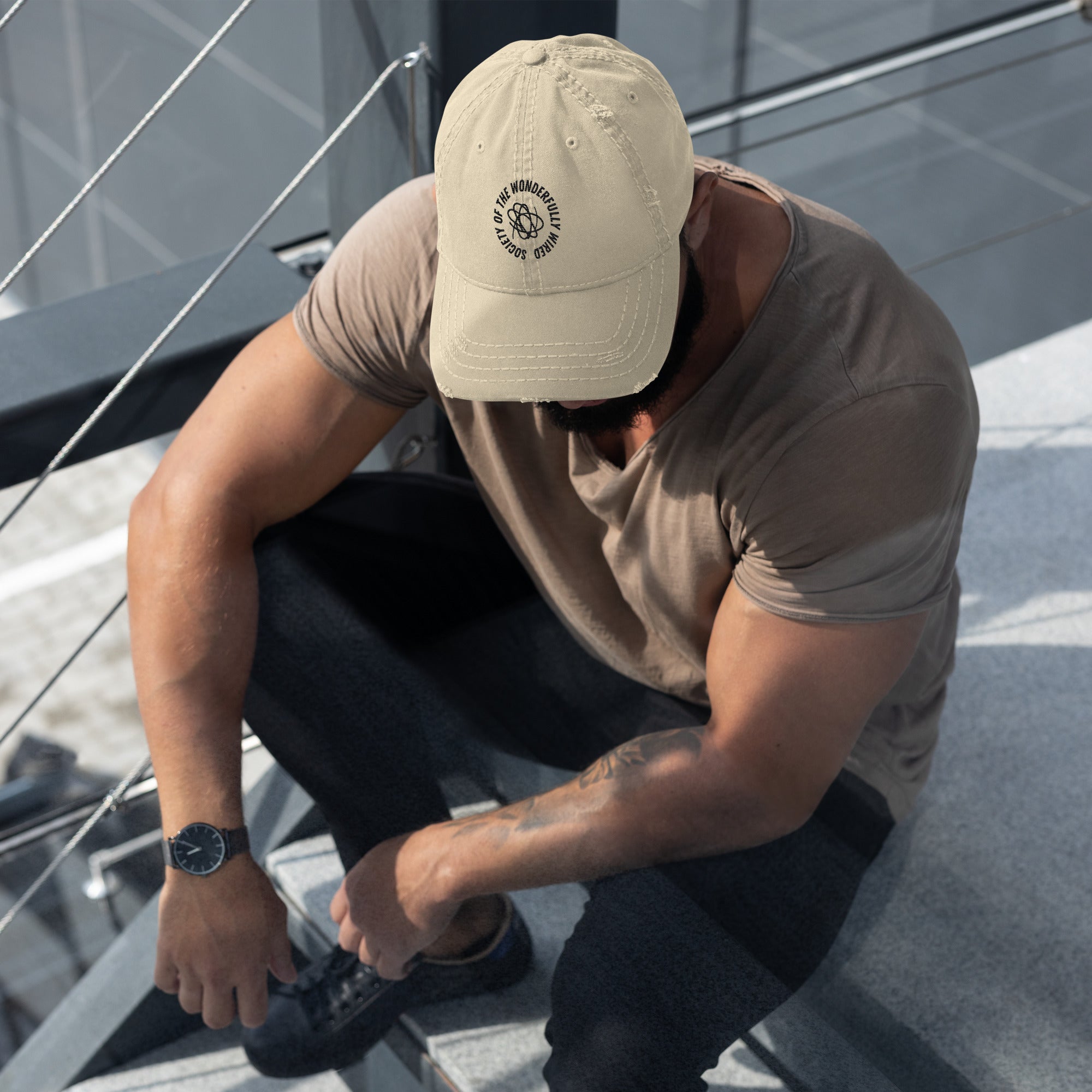 Image of a man wearing a cap with "society of the wornderfully wired" embroidered on it. Cap is beige.