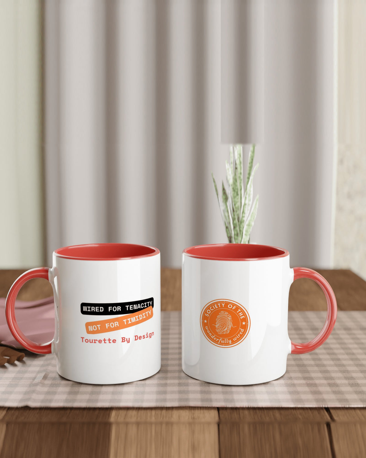 Two red-trim Tourette By Design mugs on a checked cloth, one showing Wired for Tenacity Not for Timidity, the other the orange Society of the Wonderfully Wired badge