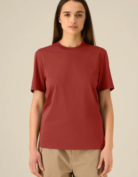 A woman modeling a short-sleeve crewneck t-shirt in a muted brick red color, paired with beige trousers, against a solid light beige background.