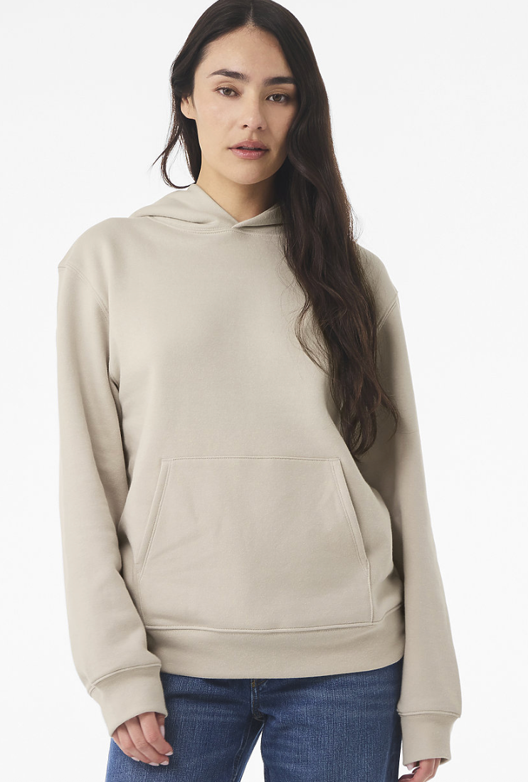 A woman modeling a light beige pullover hoodie with a front kangaroo pocket, paired with blue jeans, against a plain white studio background.