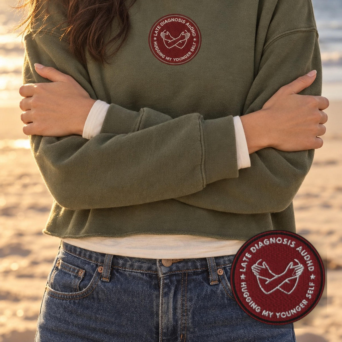 Woman on a beach with her arms wrapped around herself in an olive green crop hoodie with an embroidered late diagnosis AuDHD badge